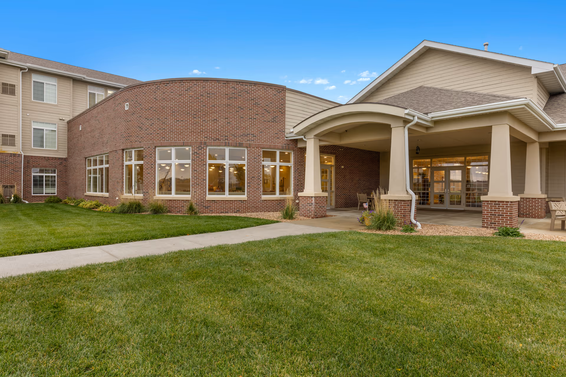 Exterior view of Whispering Creek Senior Living facility showing a well-maintained lawn, a curved brick wall with large windows, and a covered entrance supported by columns under a clear blue sky.