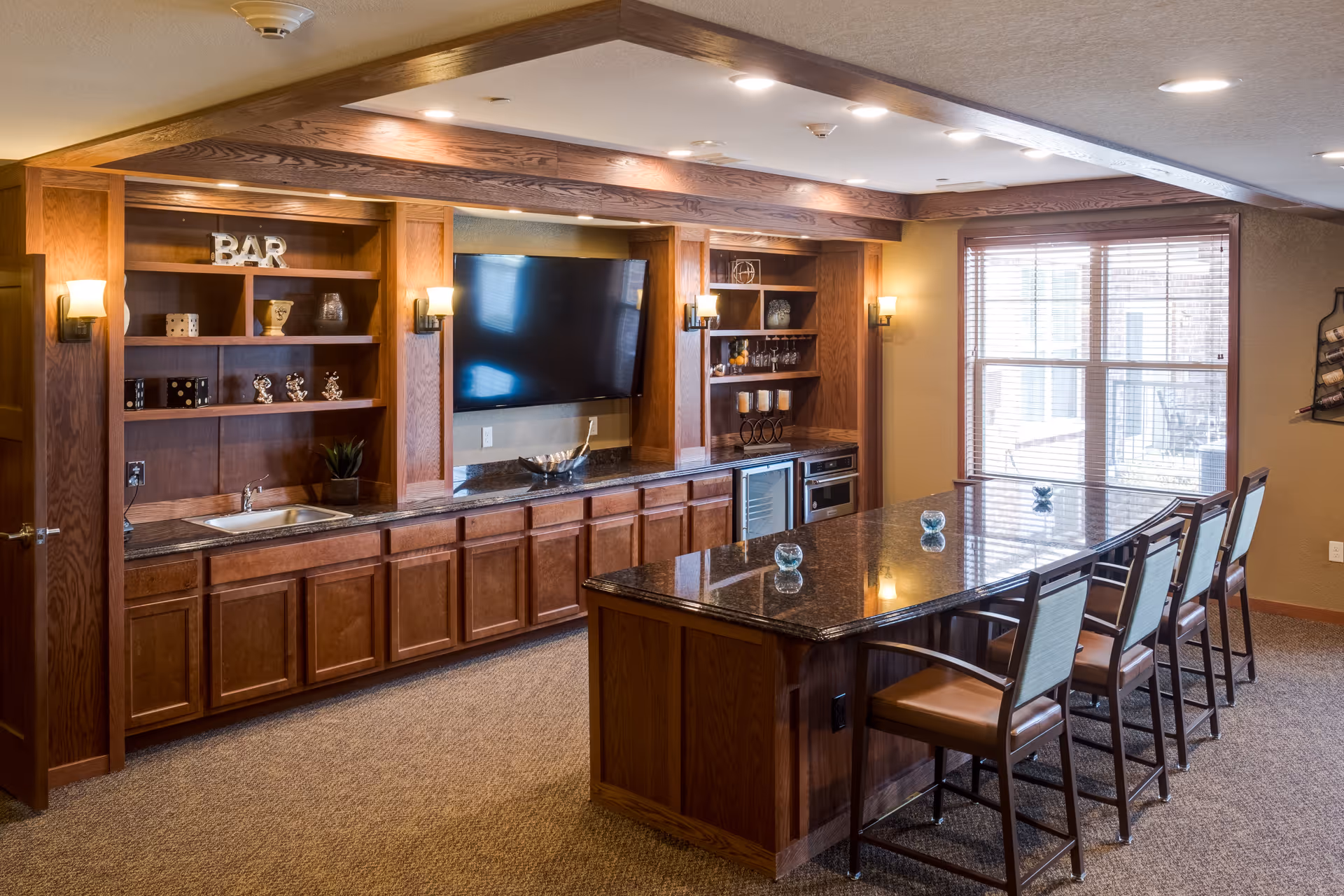 A spacious room with a long granite countertop island surrounded by six high chairs. The back wall features wooden cabinetry with shelves, a small sink, a mounted flat-screen TV, and a mini refrigerator. The room is warmly lit with wall sconces and recessed ceiling lights, and there is a large window with blinds letting in natural light.
