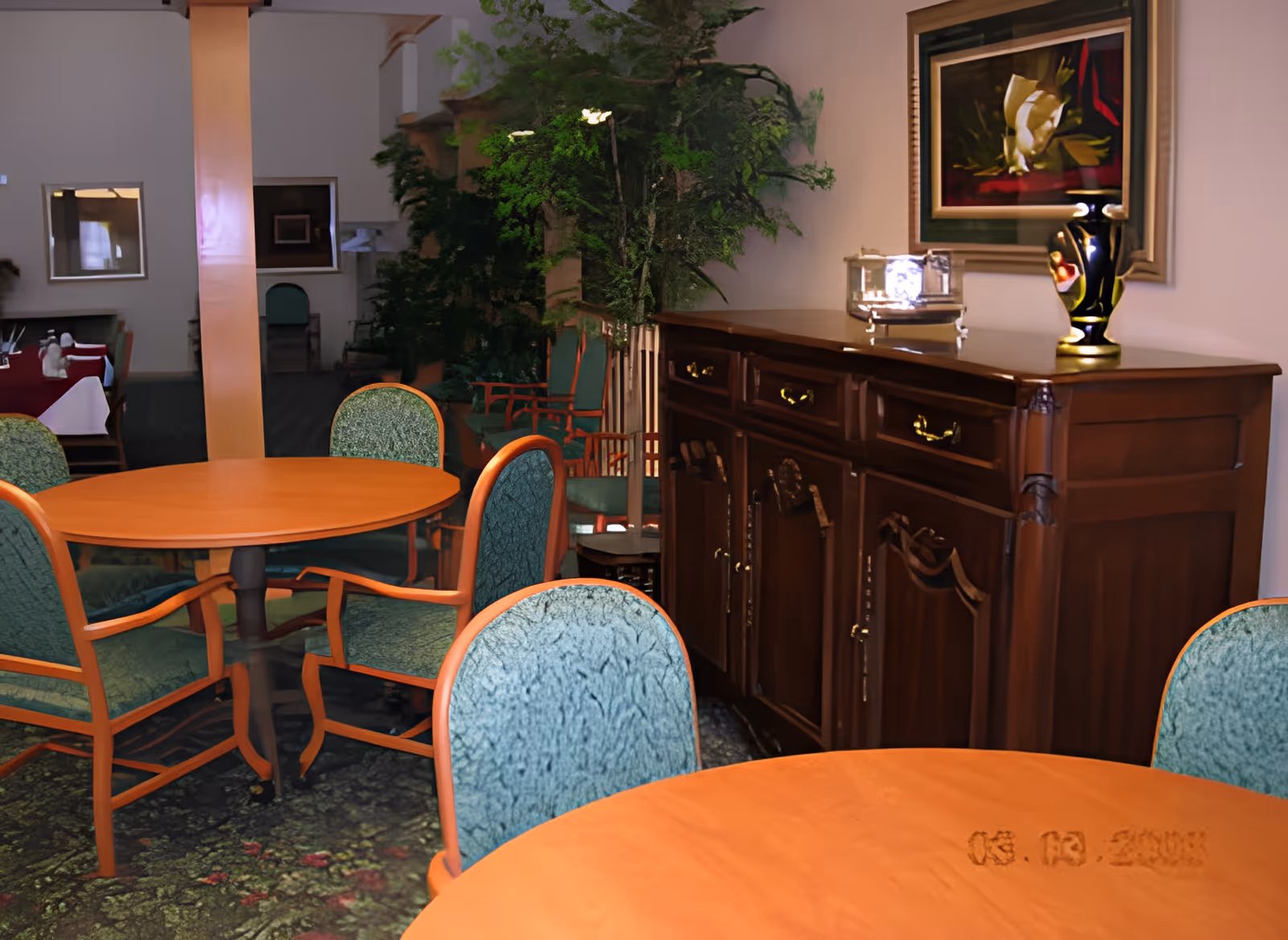 Interior view of a senior living facility dining area with round wooden tables and green upholstered chairs. A wooden sideboard with decorative items and a framed painting is visible against the wall. There are plants and additional seating in the background.