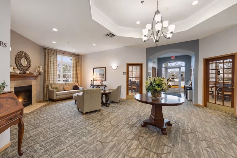 A spacious and well-lit common area in a senior living facility featuring a round wooden table with a floral arrangement in the center, comfortable seating including a sofa and armchairs near a fireplace, large windows with curtains, and double glass doors leading to other rooms.