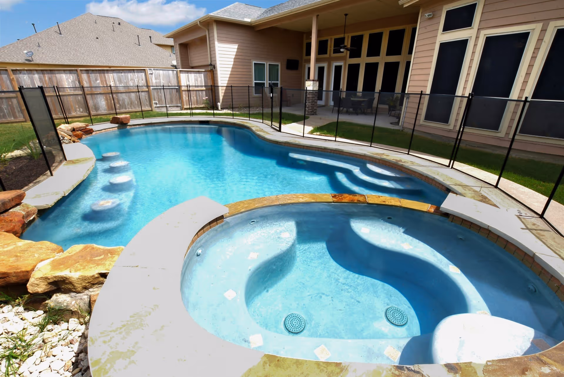 Outdoor swimming pool and attached hot tub with clear blue water, surrounded by a safety fence. The pool area is adjacent to a beige building with large windows and a covered patio with outdoor furniture. There is a wooden fence enclosing the backyard area.