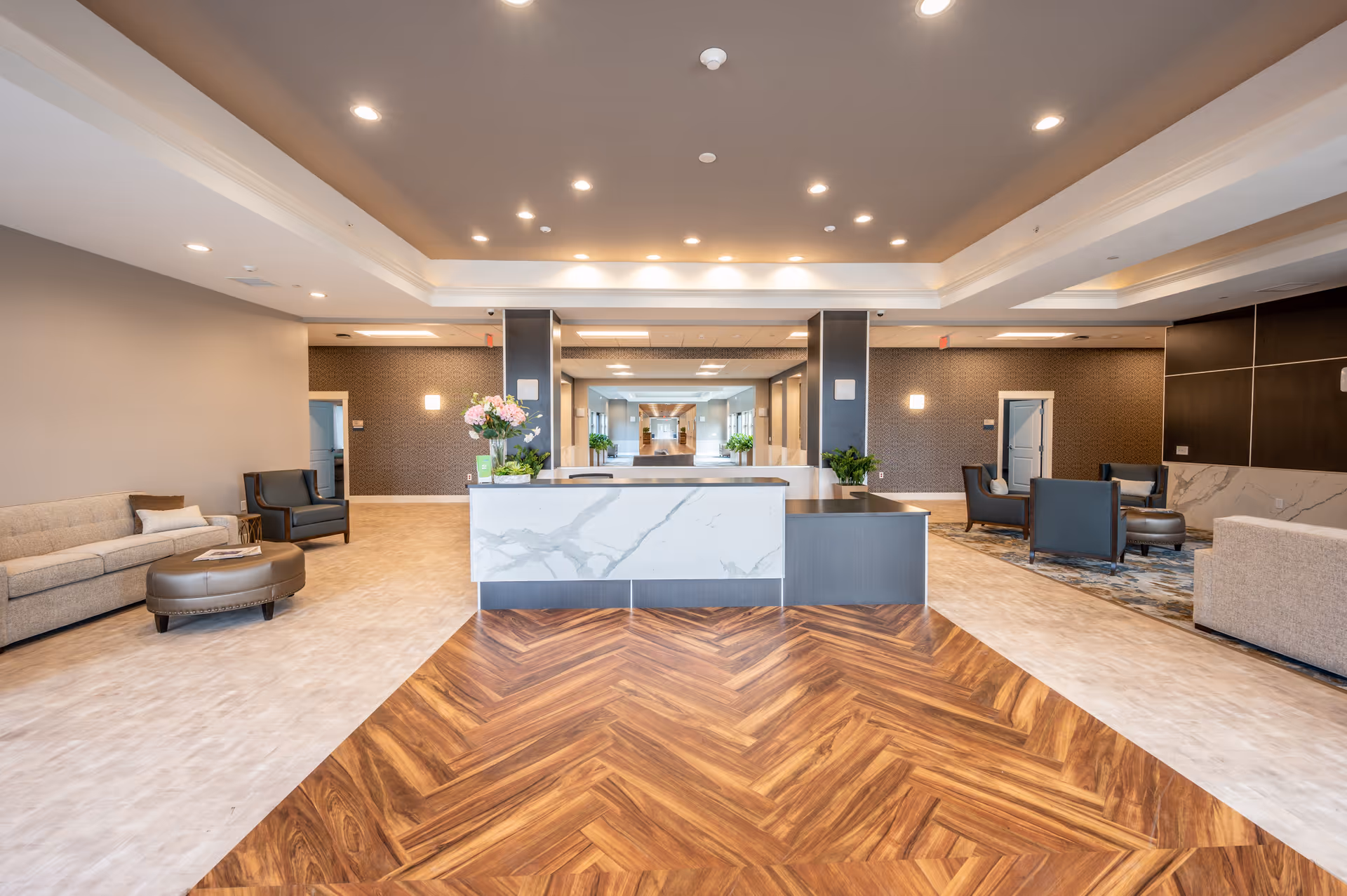Spacious and modern senior living facility lobby with a central reception desk featuring a marble front, surrounded by comfortable seating areas with sofas and armchairs. The floor has a combination of wood-patterned and light-colored tiles, and the ceiling is equipped with recessed lighting. Decorative plants and flowers add a welcoming touch.