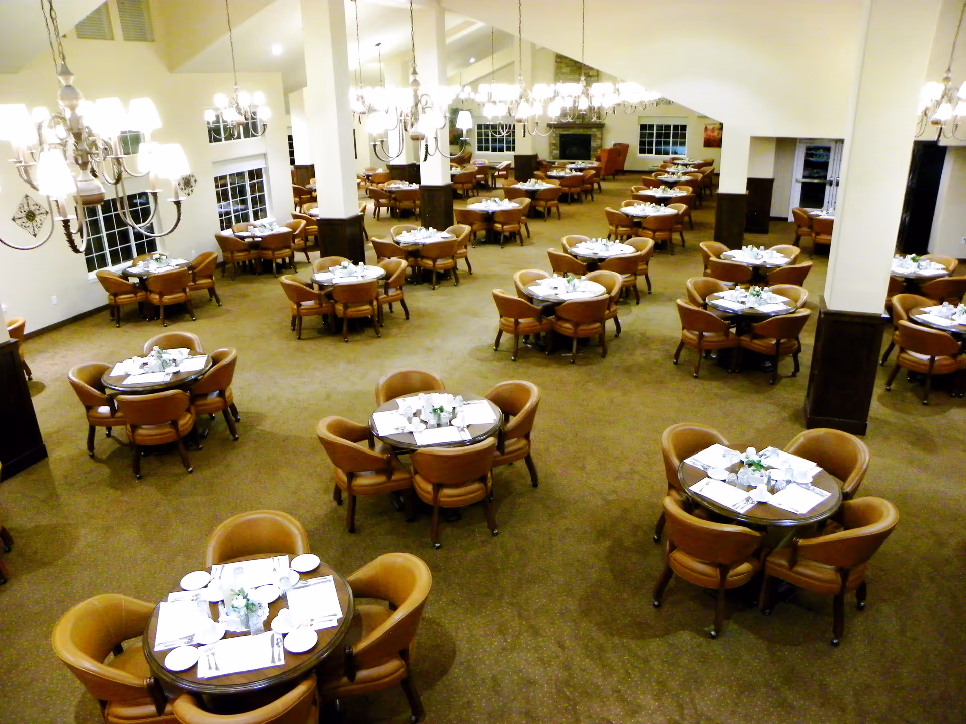 A spacious dining room with multiple round tables, each surrounded by four cushioned chairs. The tables are set with white plates, napkins, and silverware, and small floral centerpieces. The room is well-lit with chandeliers hanging from the ceiling and large windows along the walls.