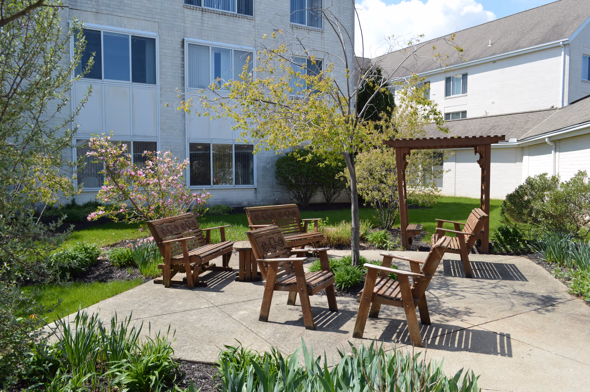 Outdoor seating area with wooden chairs and benches arranged around a small round table on a concrete patio, surrounded by green grass, flowering plants, and small trees, with a white brick building in the background under a partly cloudy sky.