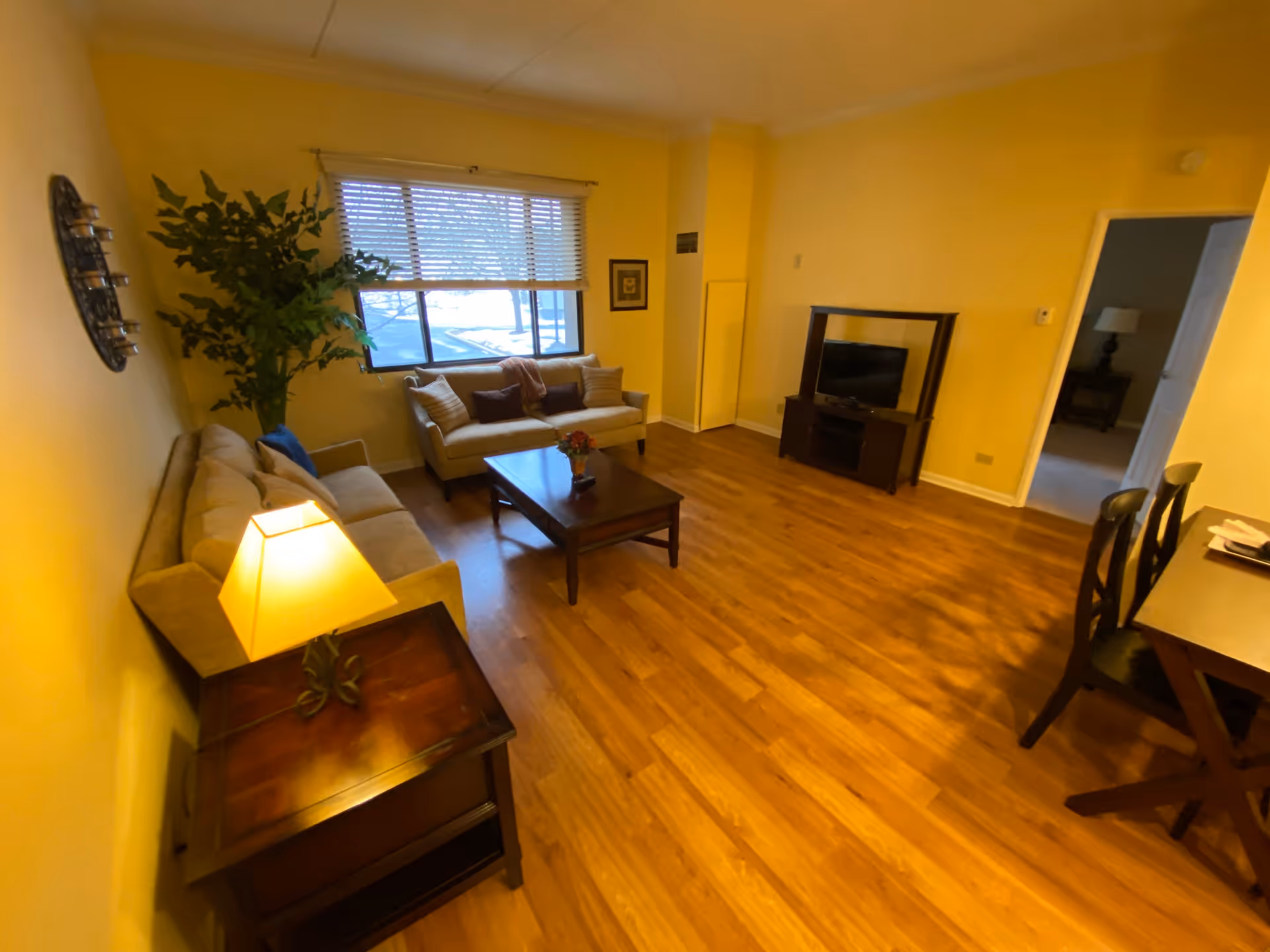 A cozy living room with wooden flooring, two beige sofas with cushions, a wooden coffee table with a small flower arrangement, a side table with a lit lamp, a large window with blinds, a TV on a stand, and a dining table with chairs. The walls are painted yellow and there is a large green plant in the corner.