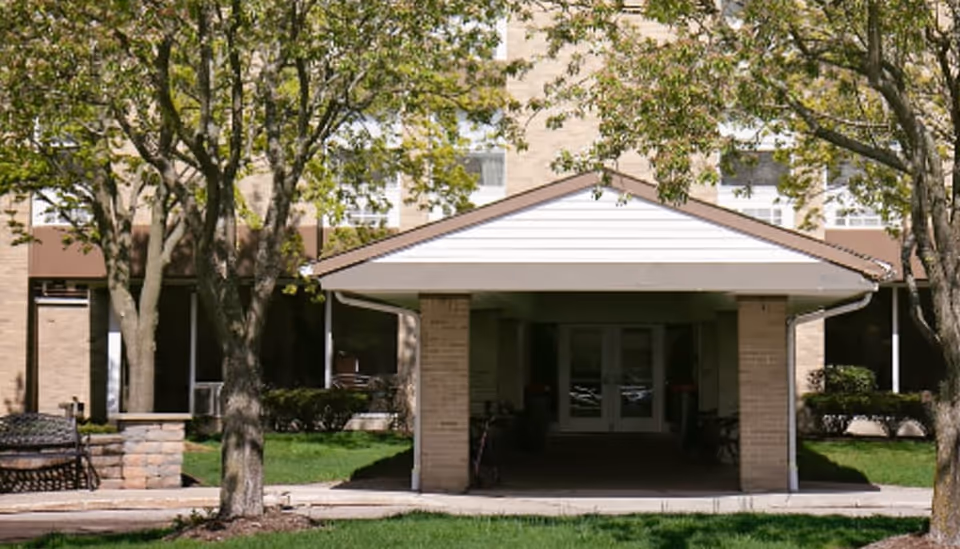 Entrance of a senior living facility named Riverside Residence with a covered porch supported by brick columns, surrounded by trees and green grass.