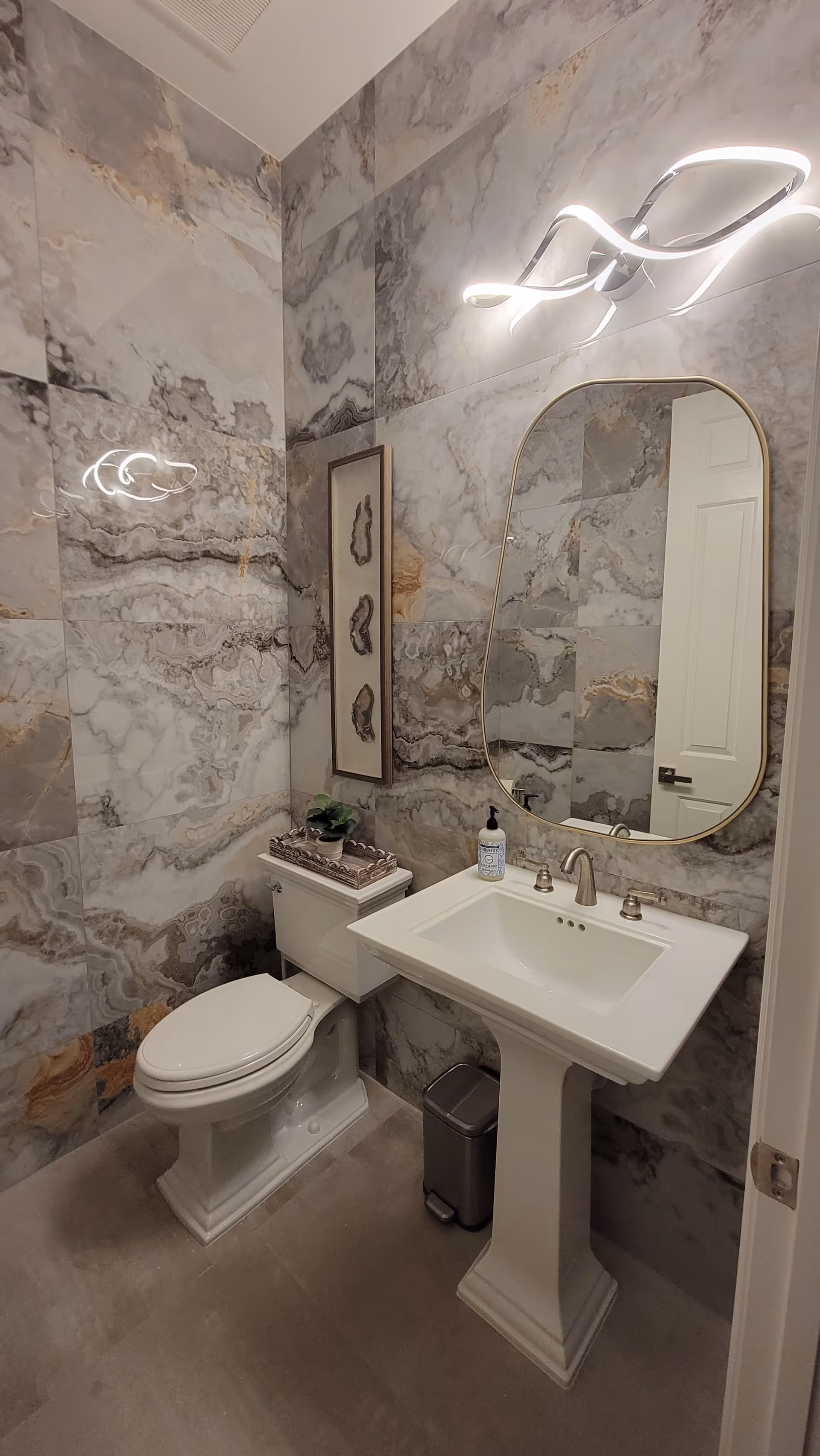 A small bathroom with marble-patterned walls, featuring a white pedestal sink with a modern faucet, a rectangular mirror with rounded edges above the sink, a toilet with a decorative tray and plant on the tank, a small trash can on the floor, and a contemporary light fixture above the mirror.
