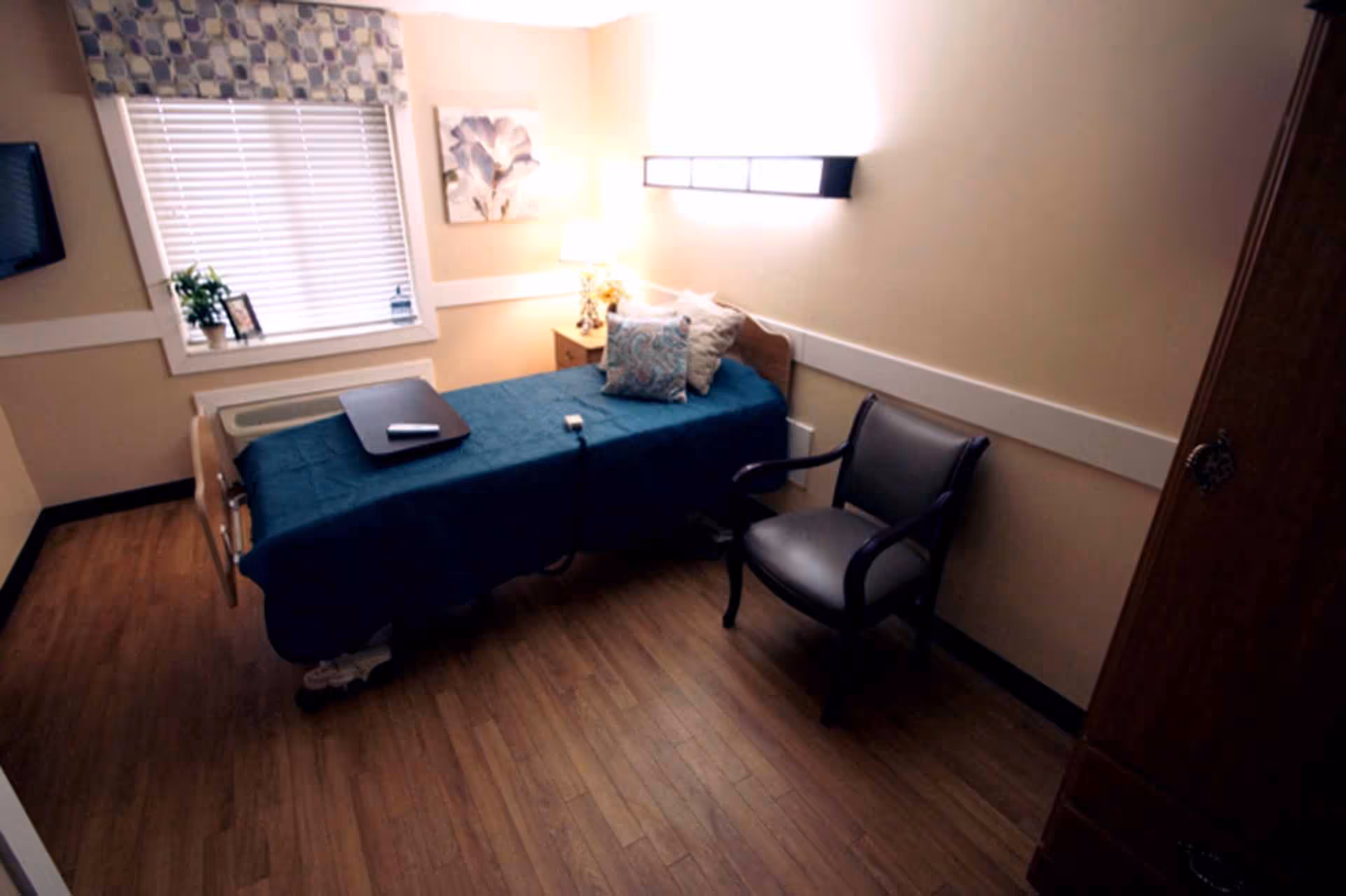 A small nursing facility bedroom with a single bed covered in a blue blanket, a bedside table with a lamp, a window with blinds and a patterned valance, a wall-mounted light fixture, a cushioned armchair, and wooden flooring.