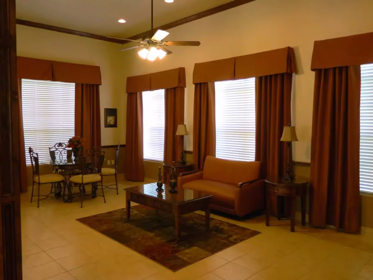 Sunlit common room with an upholstered loveseat, coffee table and small dining set in front of tall windows with brown drapes.