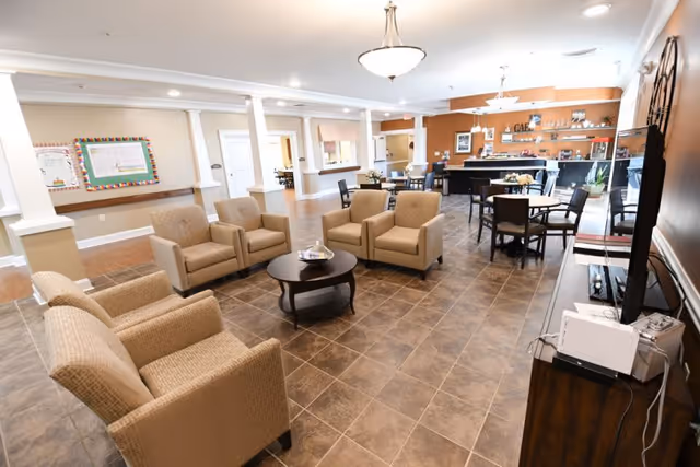 Spacious senior living common area with upholstered armchairs around a coffee table, dining tables, and a café counter in the background.