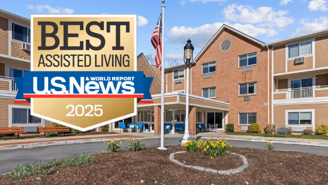 Exterior view of a multi-story assisted living facility with a circular driveway, landscaped flower beds, an American flag on a flagpole, and outdoor seating visible on the porch under a blue sky with some clouds.