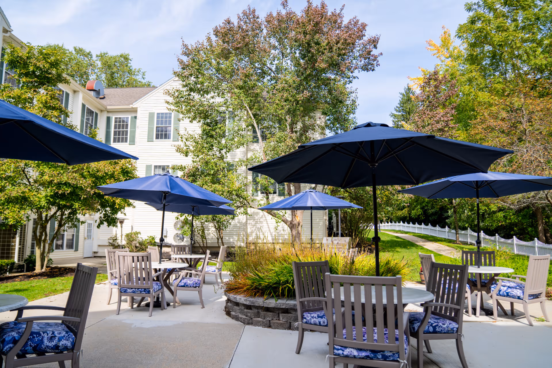 Outdoor patio with round tables, chairs, and blue umbrellas in front of a light-colored multi-story building.