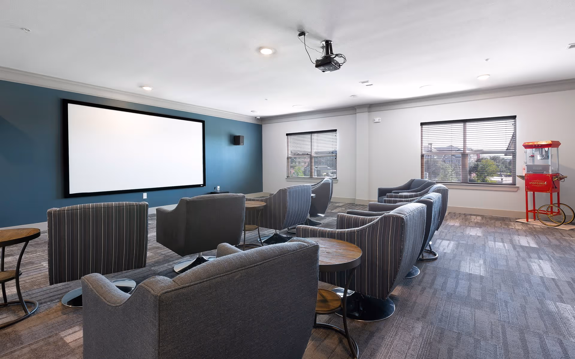 A modern media room with a large projector screen on a blue accent wall, multiple comfortable gray and striped armchairs arranged in rows facing the screen, small round wooden side tables between the chairs, two windows with blinds letting in natural light, and a red popcorn machine in the corner.