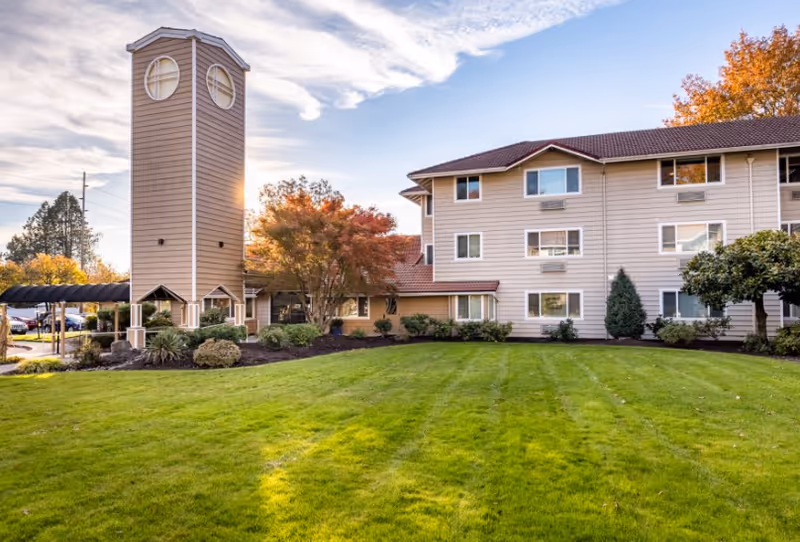Exterior view of Van Mall Retirement Vancouver facility showing a well-maintained green lawn, a tall clock tower structure, and a multi-story beige building with several windows. Trees with autumn foliage are visible around the building under a partly cloudy sky.
