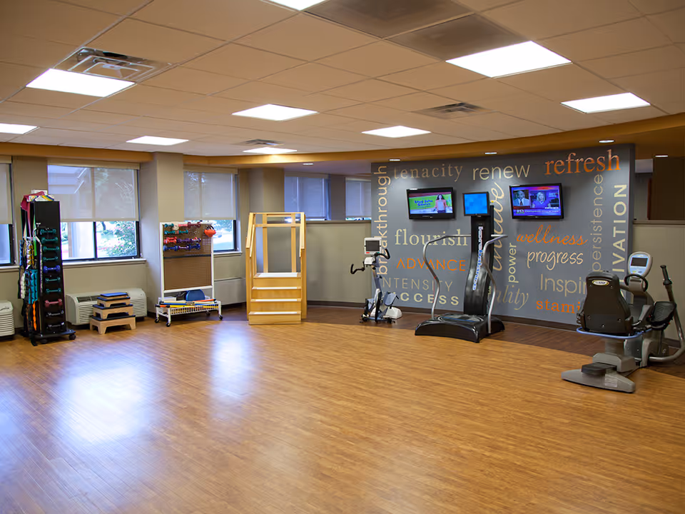 A spacious exercise room with wooden flooring and large windows with blinds. The room contains various fitness equipment including dumbbells, step platforms, a stationary bike, and a treadmill. The back wall features motivational words such as 'tenacity', 'renew', 'refresh', and 'wellness' along with three mounted flat-screen TVs.
