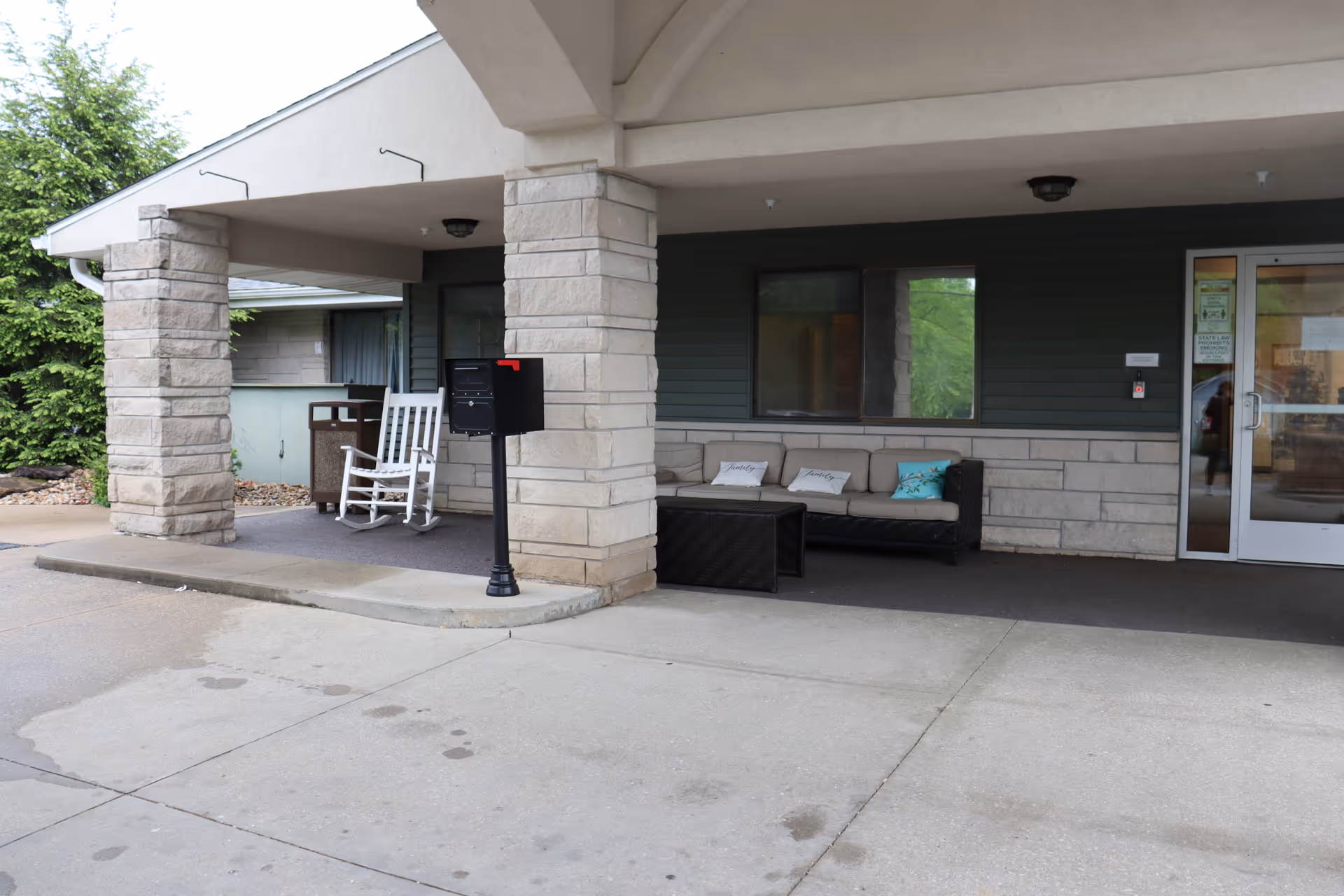 Covered entrance area of a building with stone pillars, a white rocking chair, a black mailbox on a post, and a black outdoor sofa with beige cushions and decorative pillows. There is a glass door entrance on the right side and greenery visible in the background.
