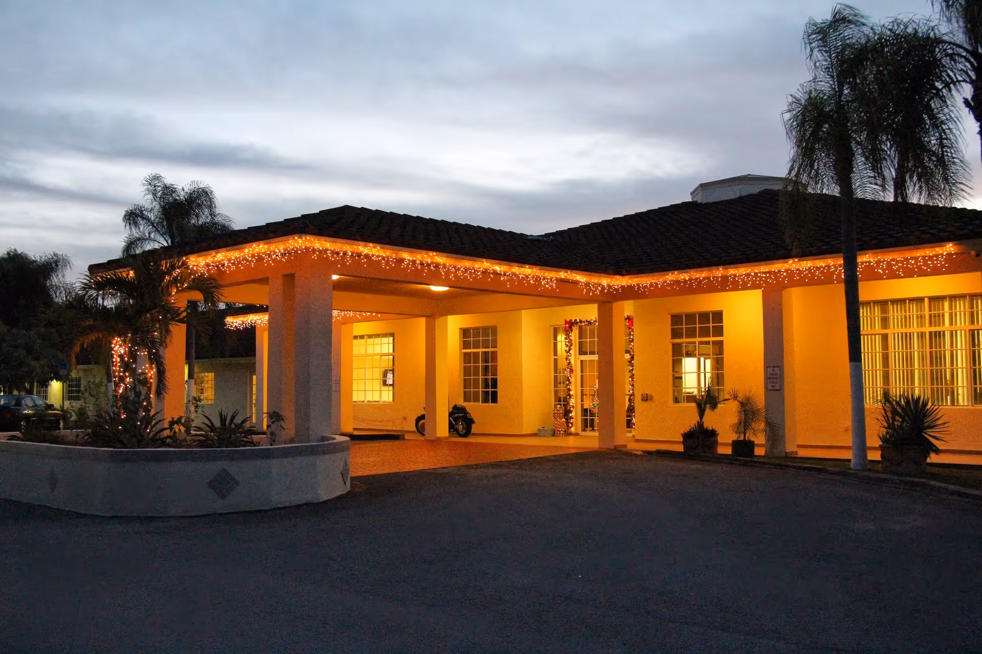 Warmly lit entrance canopy of an assisted living building decorated with string lights and palm trees at dusk.