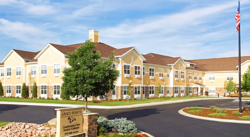Exterior view of Grace Pointe Senior Care Community, a large yellow two-story building with white trim and a brown roof. The building is surrounded by a well-maintained lawn, trees, and a paved driveway. A sign in front reads 'Grace Pointe Continuing Care Senior Campus 1919 68th Avenue'. An American flag is visible on a flagpole to the right.