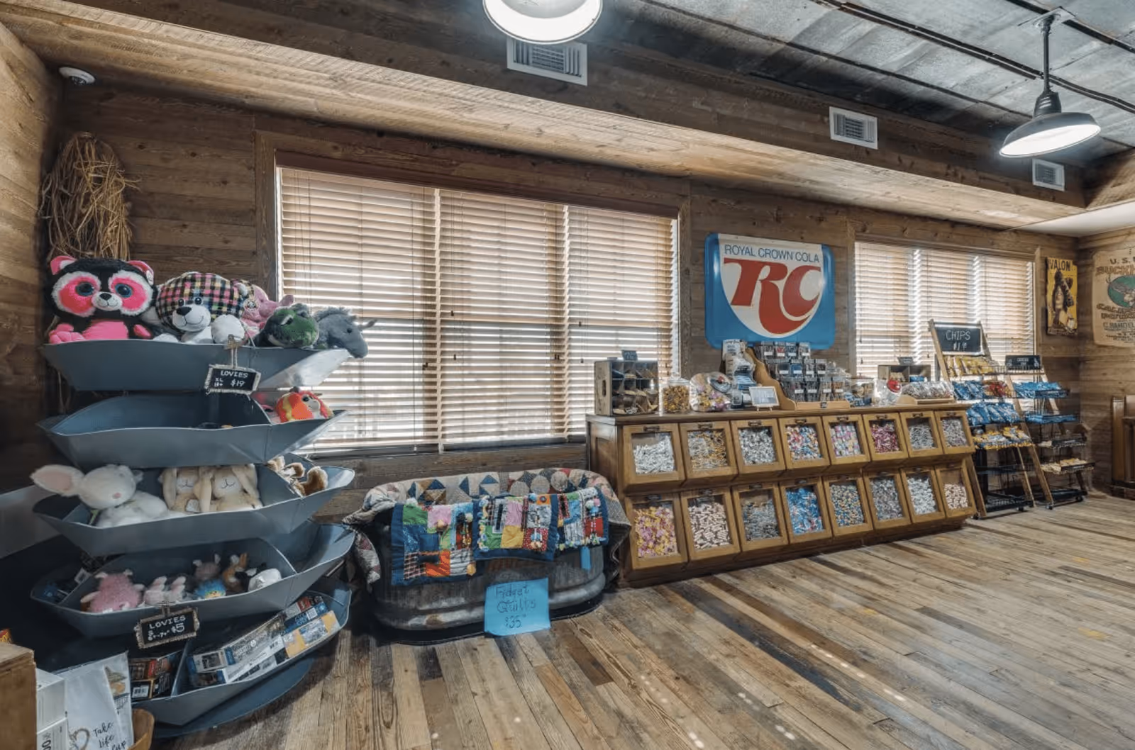 Interior of a rustic store or gift shop with wooden walls and floor. On the left, there are metal tiered shelves holding stuffed animals and toys. In the center, a metal tub contains colorful folded quilts with a sign reading 'Fidget Quilts $35'. On the right, wooden bins display various packaged snacks and candies under a Royal Crown Cola sign. The room is lit by ceiling lights and natural light coming through the windows with blinds.
