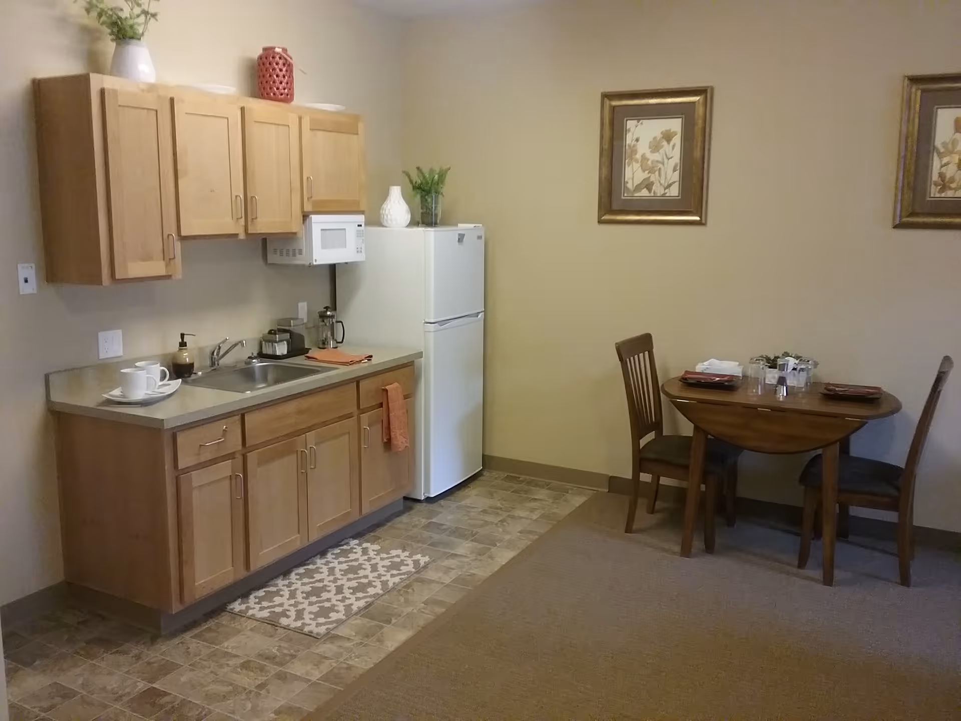 A small kitchen area with wooden cabinets, a countertop with a sink, a white microwave, and a white refrigerator. To the right, there is a round wooden dining table with two chairs, set with plates, glasses, and napkins. The walls are beige with two framed floral pictures hanging above the dining table.