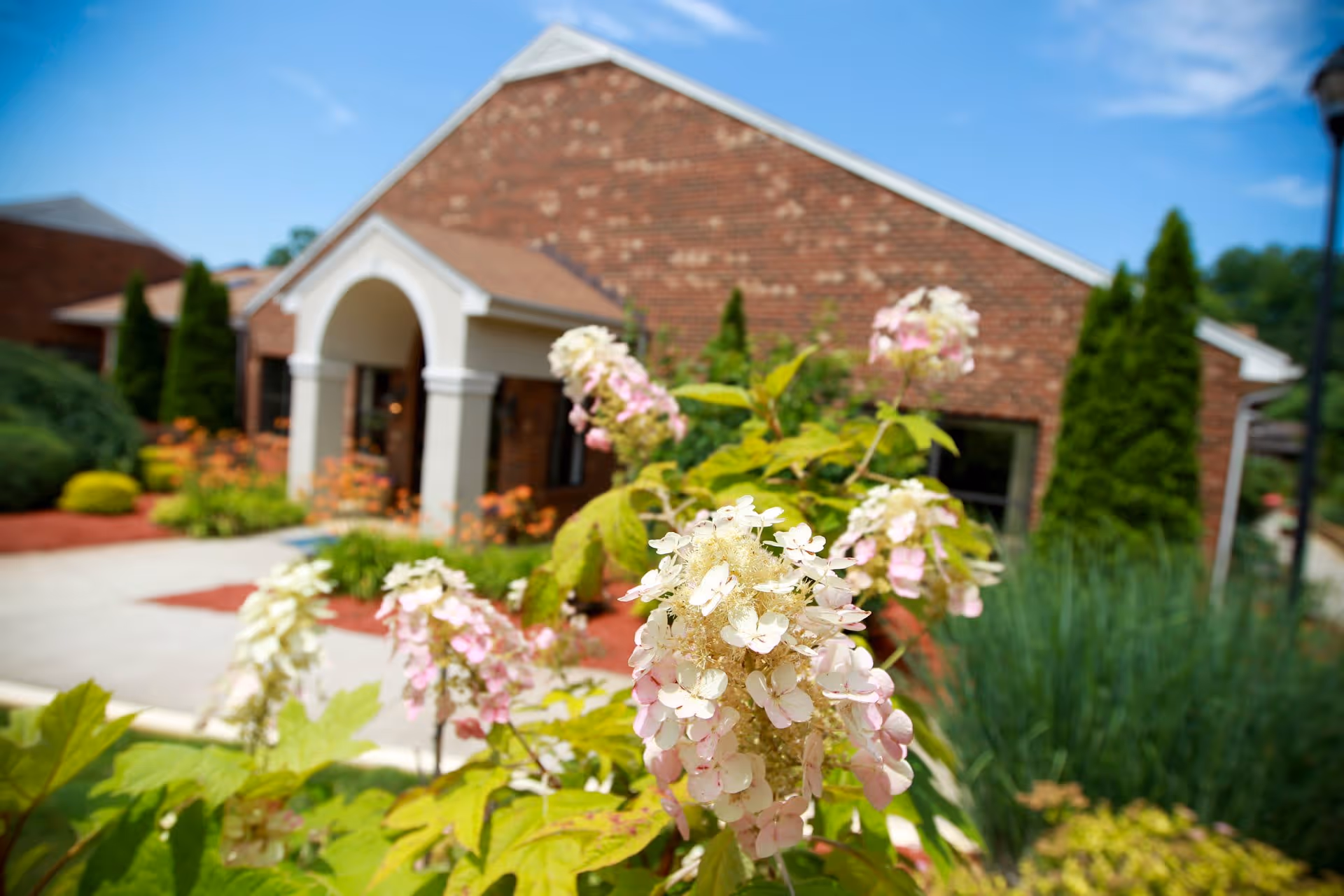 Close-up of pink and white flowers in front of a brick building with an arched entrance, surrounded by greenery and landscaping under a blue sky.