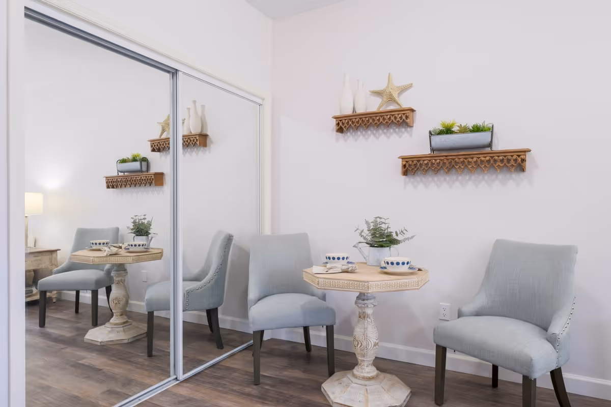 A small dining area with a round wooden table set for two with cups and napkins. Two light blue upholstered chairs are placed around the table. The wall behind has two decorative wooden shelves with vases, a starfish, and a planter with greenery. A large mirrored sliding closet door reflects the scene, and a wooden floor lamp is visible in the reflection.