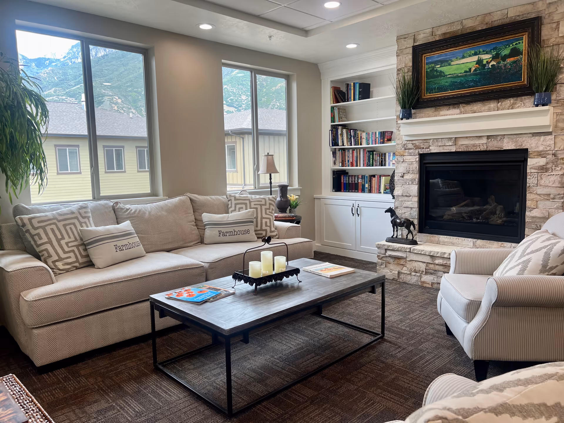 A cozy senior living facility common area with a beige sofa adorned with patterned and 'Farmhouse' pillows, a matching armchair, a wooden coffee table with candles and magazines, a stone fireplace with a landscape painting above it, built-in white bookshelves filled with books, and large windows showing a view of neighboring buildings and mountains.