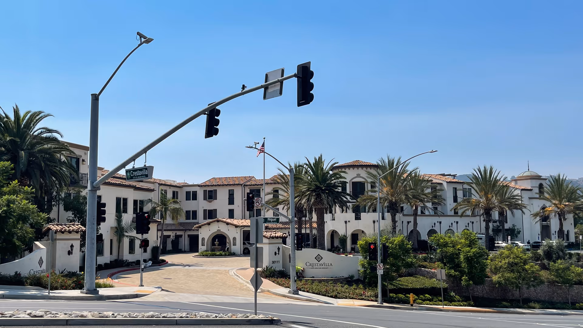 Exterior view of the Crestavilla senior living facility showing a large, multi-story building with Spanish-style architecture, palm trees, and a driveway entrance. Traffic lights and street signs for Crestavilla and Niguel are visible in the foreground under a clear blue sky.