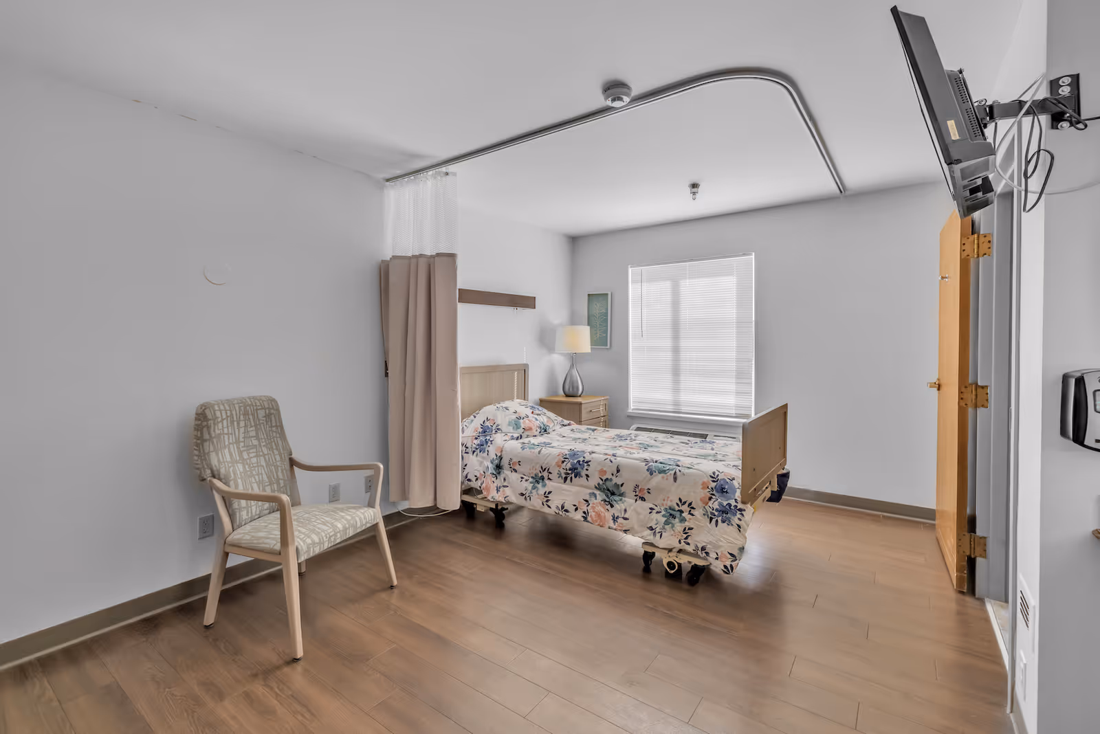 A simple nursing home patient room with a single bed on wheels, bedside table and lamp, privacy curtain, chair, and a wall-mounted TV.