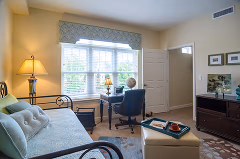 Sunlit living room with a daybed, desk and chair by a large window, an ottoman with a tray, and a media cabinet.