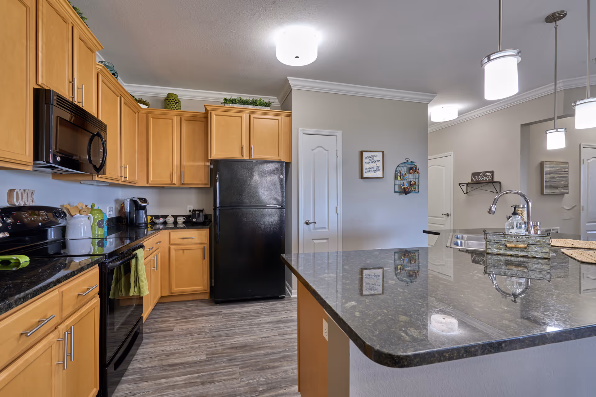 Modern kitchen with wooden cabinets, black appliances including a refrigerator, stove, and microwave. The kitchen features a large granite countertop island with a sink and a faucet. The floor is wood-patterned, and the walls are painted light gray. There are decorative items on top of the cabinets and on the walls, and three pendant lights hang above the island.