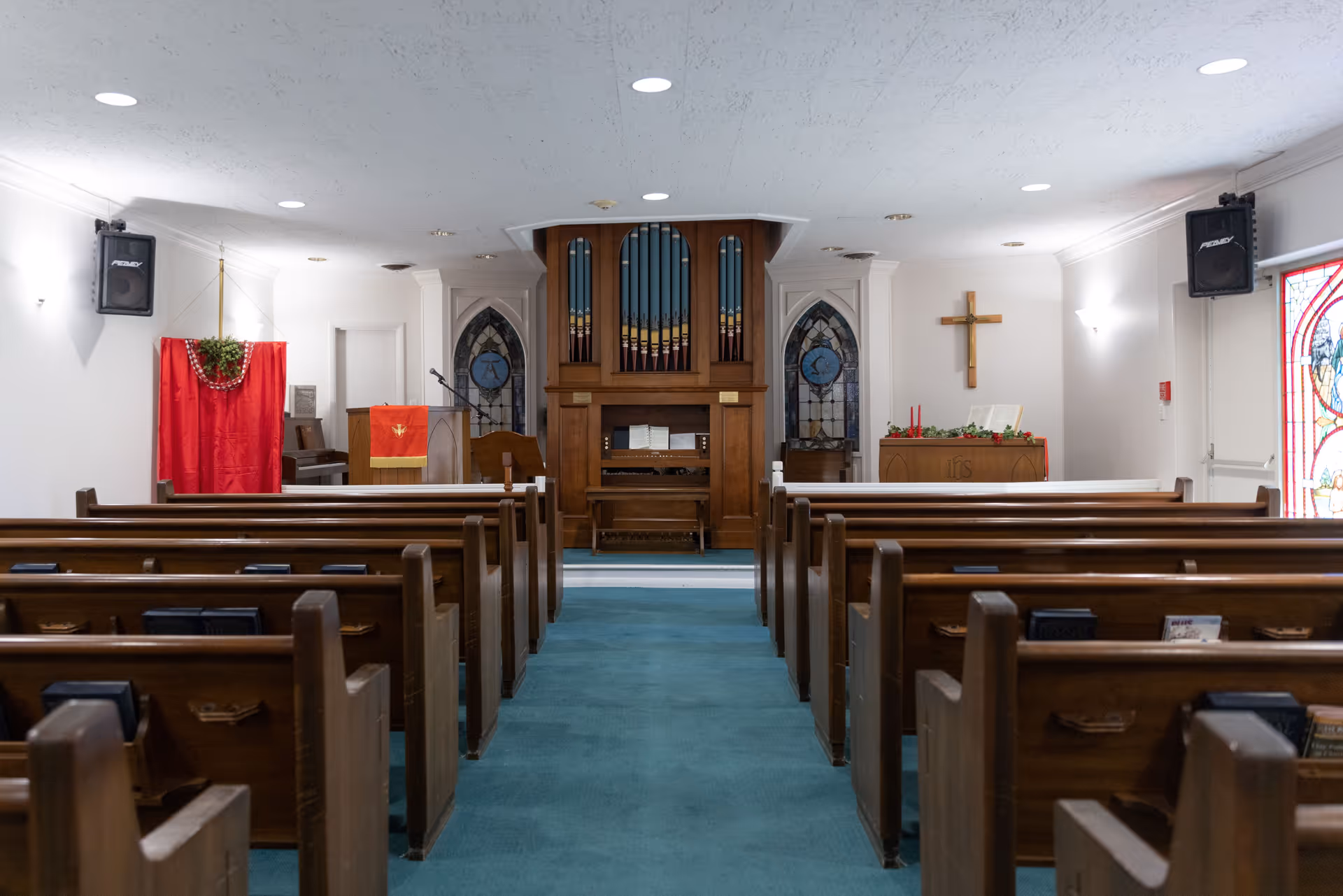 Interior view of a chapel with wooden pews arranged in rows facing a pipe organ at the front. The chapel has stained glass windows, a wooden cross on the wall, and religious decorations including red cloths and candles.