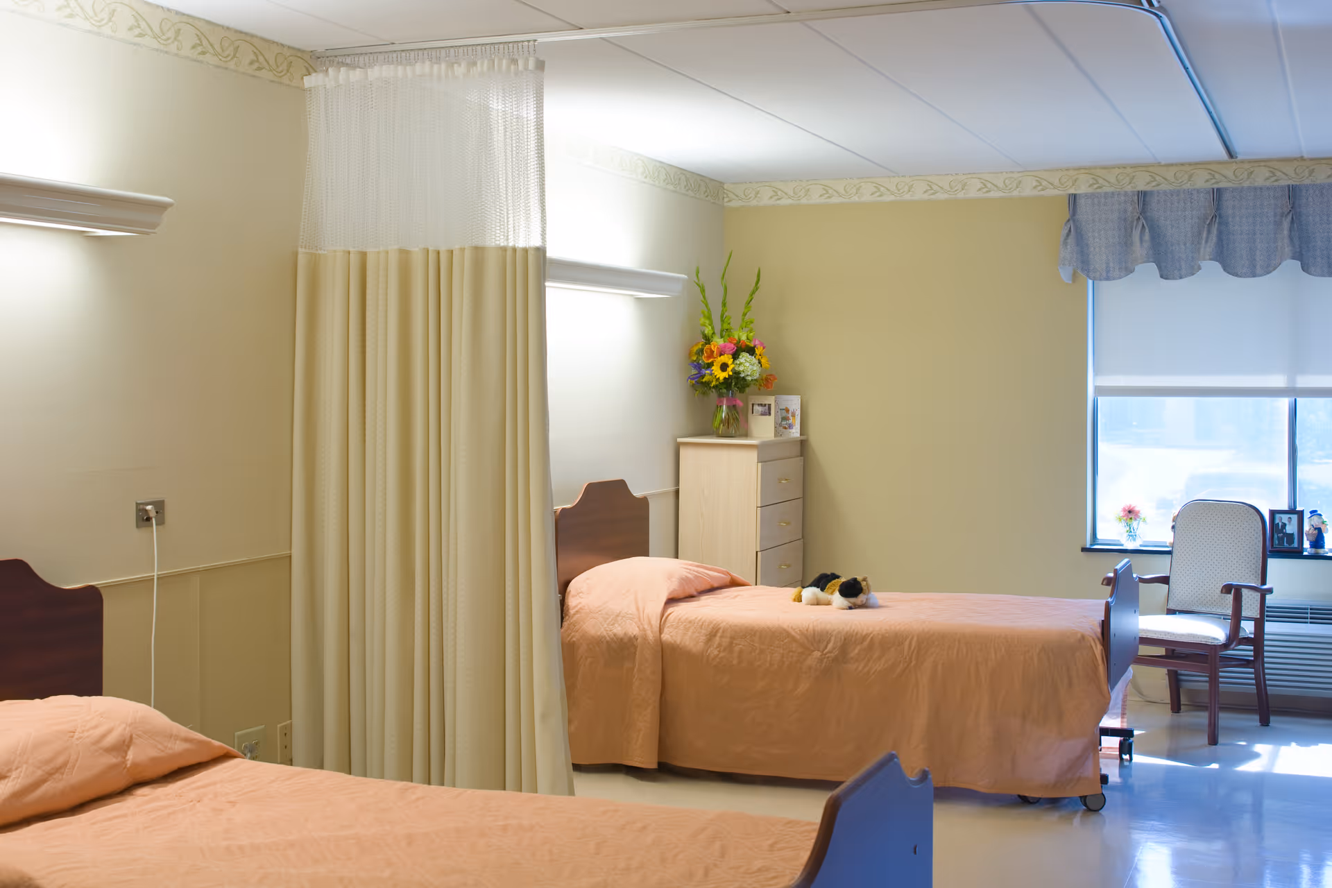 A shared bedroom in a senior living facility with two single beds covered in peach-colored bedspreads. A privacy curtain hangs between the beds. There is a wooden dresser with a vase of colorful flowers and framed photos on top. A window with a blue valance and a white roller shade lets in natural light. A wooden chair with a cushioned seat is placed near the window.