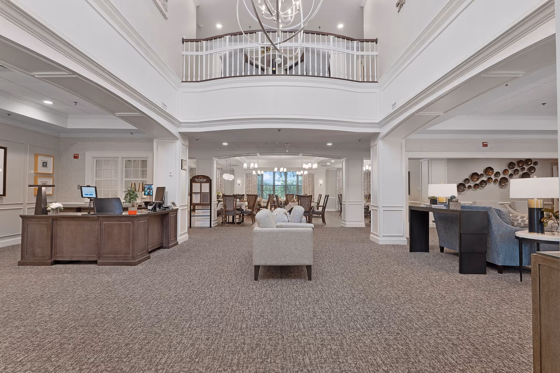 Spacious and well-lit interior common area of a senior living facility featuring a reception desk on the left, a seating area with sofas in the center, and a dining area with tables and chairs in the background. The space has high ceilings with a balcony railing above and elegant light fixtures.