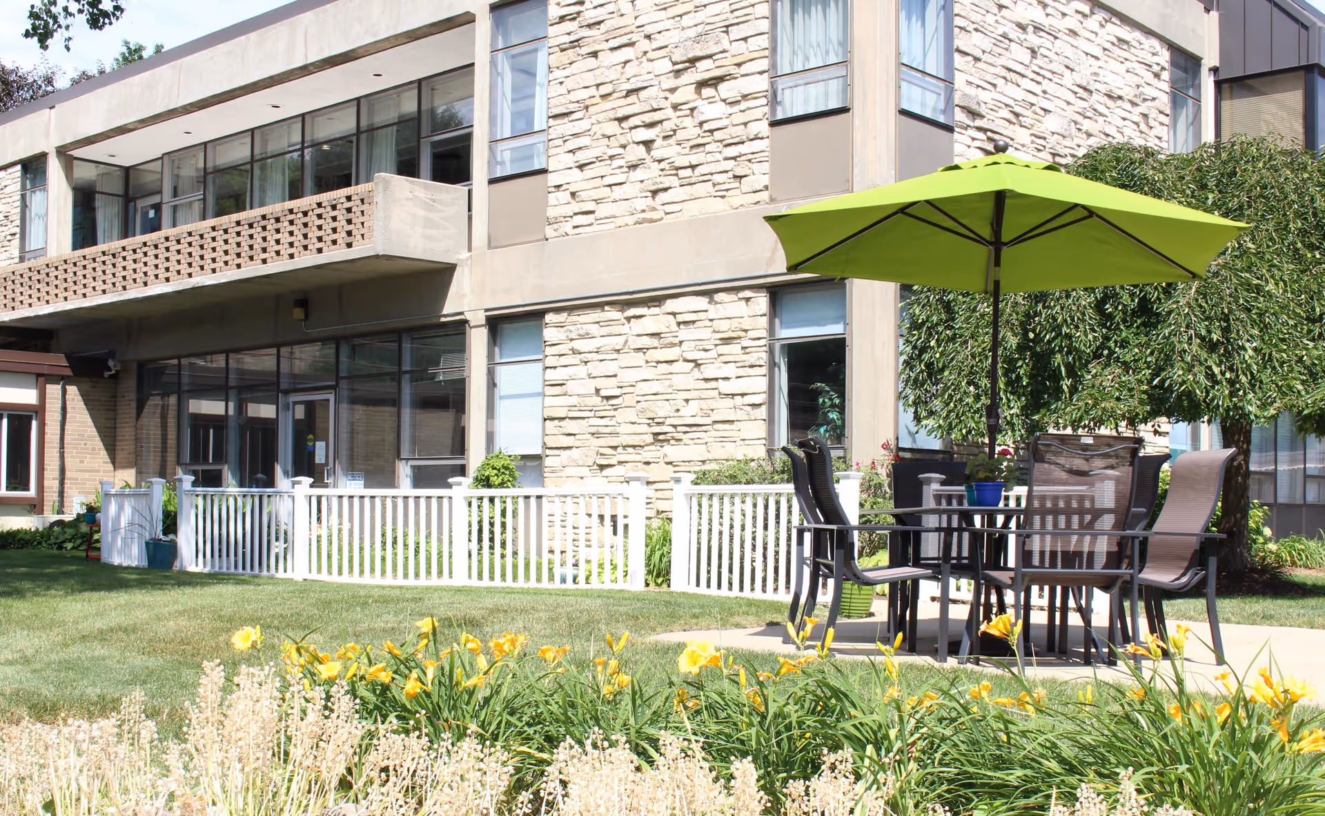 Outdoor patio area at a senior living facility with a round table, four chairs, and a large green umbrella. The patio is surrounded by green grass, yellow flowers, and shrubs. The building exterior features stone and brick walls with large windows.