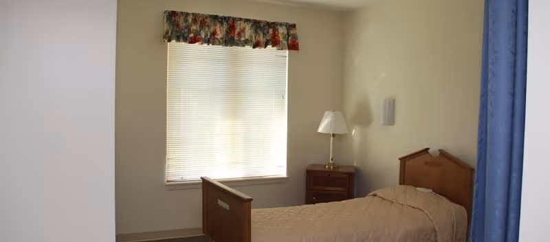 A modest bedroom with a single wooden bed, nightstand and lamp beside a window with blinds and a floral valance.