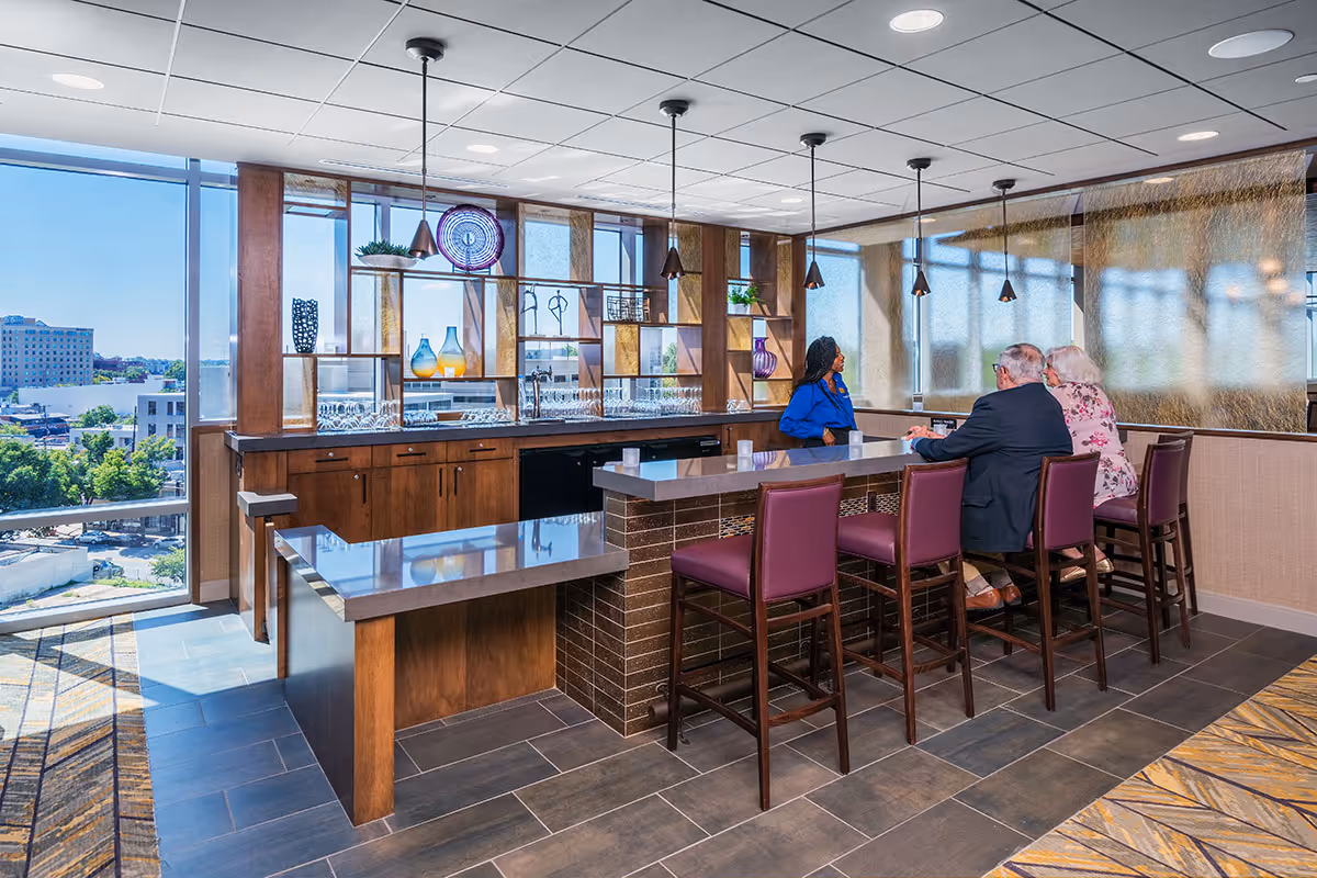 Modern communal bar area with a long counter, purple barstools, shelving with decorative glassware, large windows with a city view, and three people seated at the counter.