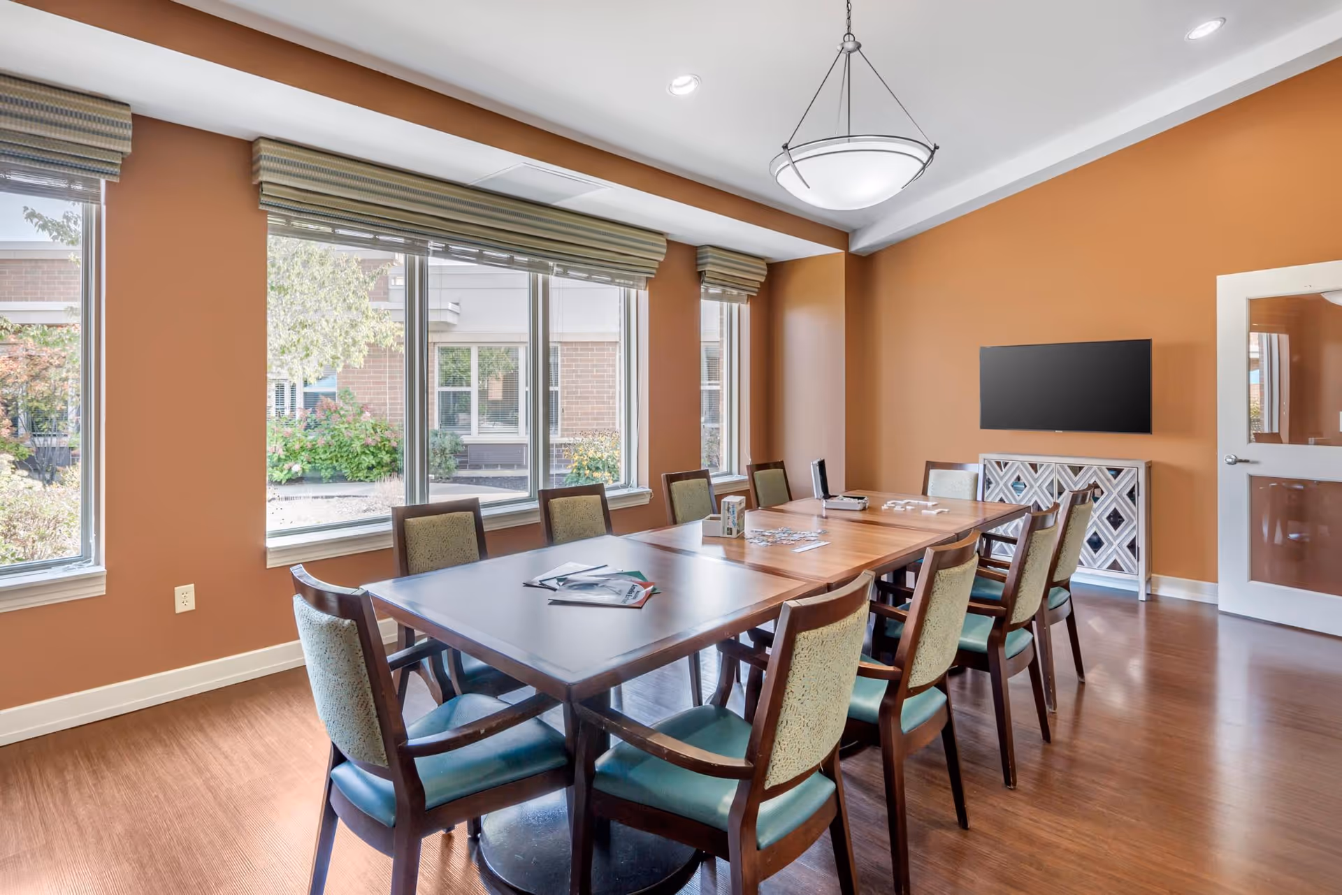 A well-lit meeting or dining room with a long wooden table surrounded by ten chairs with green cushions. The room has large windows with beige blinds, allowing natural light to brighten the space. The walls are painted a warm brown color, and there is a modern light fixture hanging from the ceiling. A flat-screen TV is mounted on the wall above a decorative white cabinet, and a door with a frosted glass panel is visible on the right side.
