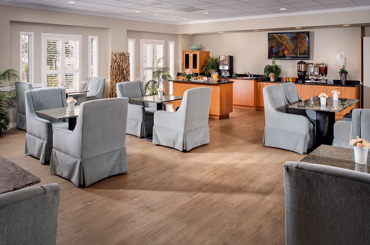 Bright communal dining area with upholstered armchairs, tables, plants, and a buffet/beverage counter.