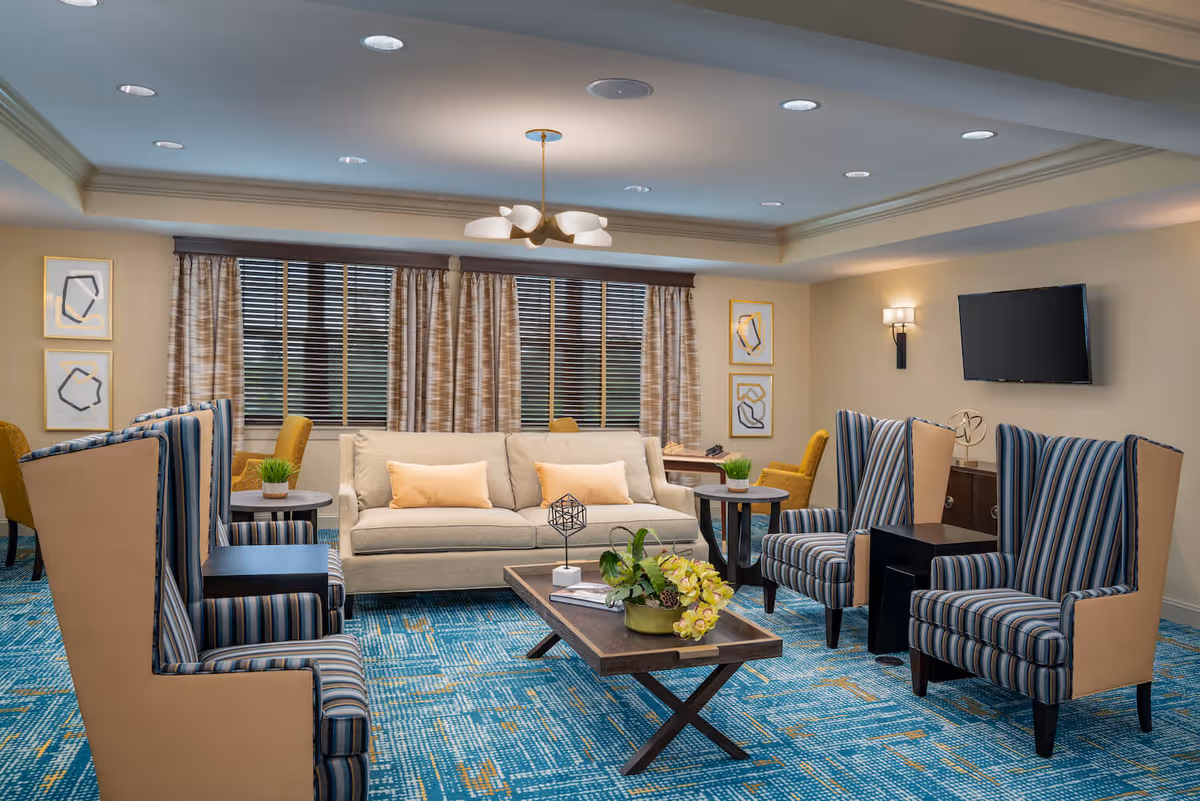 A well-lit living room with a beige sofa adorned with two light yellow pillows, four striped high-back armchairs, a wooden coffee table with a floral arrangement, and side tables with small green plants. The room features a blue patterned carpet, large windows with beige curtains, abstract wall art, and a wall-mounted flat-screen TV.