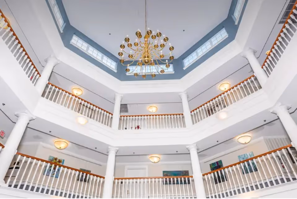 Interior view of a multi-level atrium with white railings and columns, featuring a large gold chandelier hanging from a blue and white ceiling with windows.