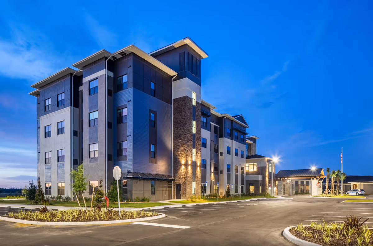 Exterior view of a modern multi-story senior living facility building during twilight with parking spaces and landscaped areas in front.