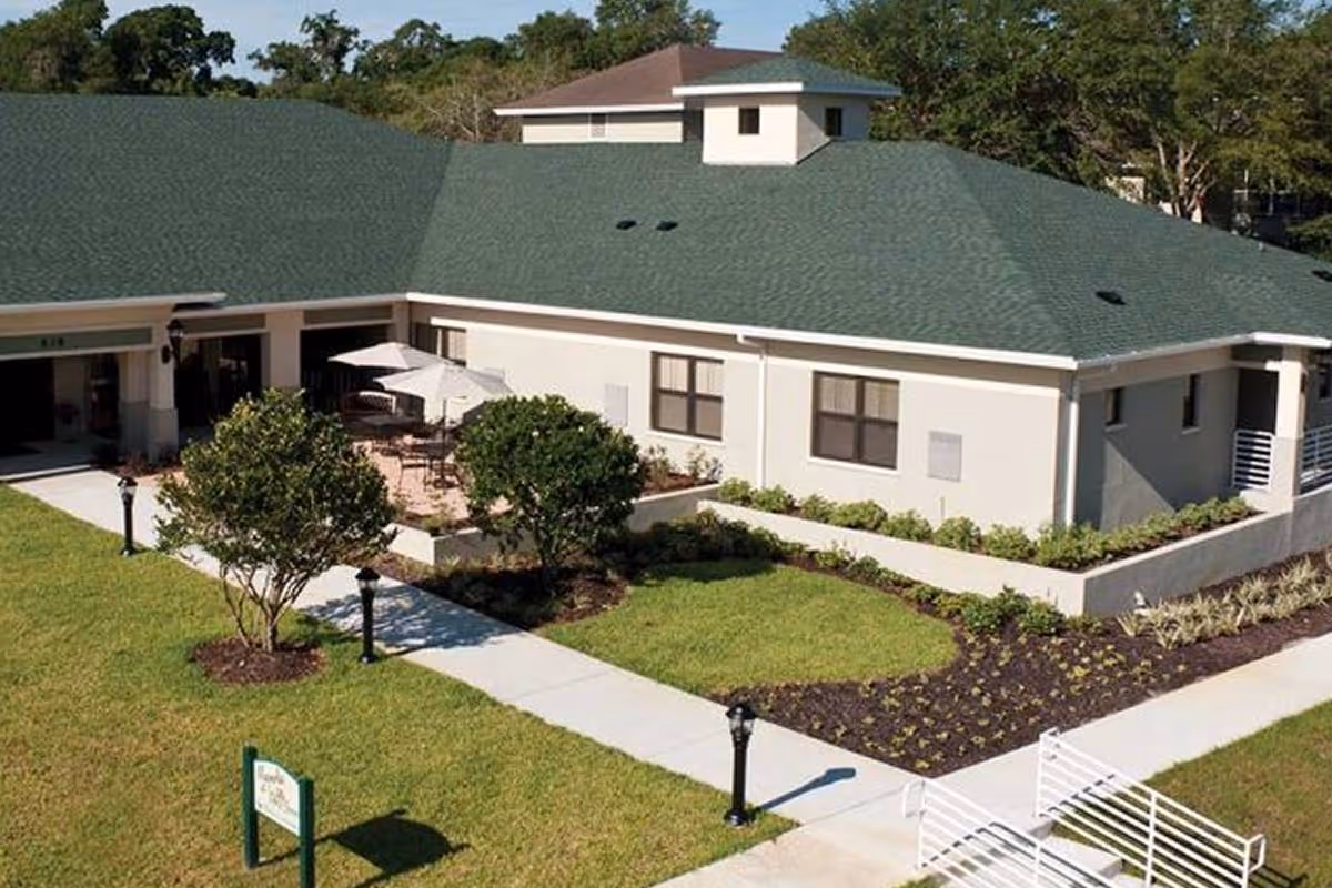 Exterior view of a single-story building with a green roof and beige walls, surrounded by well-maintained landscaping including bushes, grass, and small trees. There is a paved walkway leading to a patio area with tables and umbrellas. The sky is clear and trees are visible in the background.