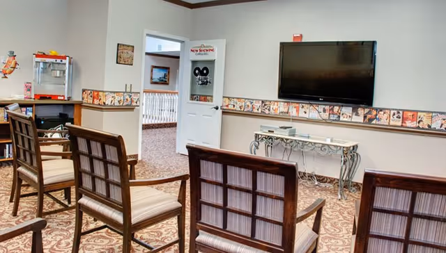 Chairs arranged facing a wall-mounted TV in a senior living common room with a popcorn machine and console table.