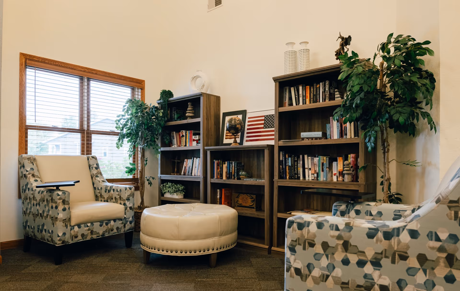 A cozy lounge area with patterned armchairs, a round ottoman, bookshelves and plants by a window.