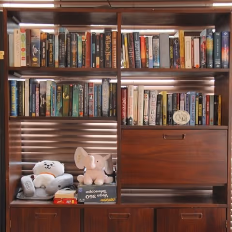 Wooden bookshelf filled with various books, two stuffed animals including a white bear and a pink elephant, and a few board games on the lower shelves.