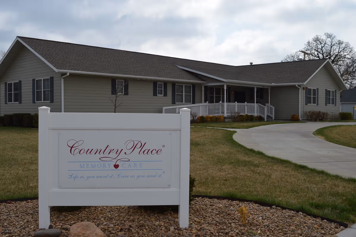 Exterior view of a single-story senior living facility building with beige siding and a dark roof. In front of the building is a white sign that reads 'Country Place Memory Care Life as you want it... Care as you need it.' The building has a porch with white railings and a curved concrete driveway leading to the entrance. The surrounding area has grass and some landscaping with rocks and bushes.