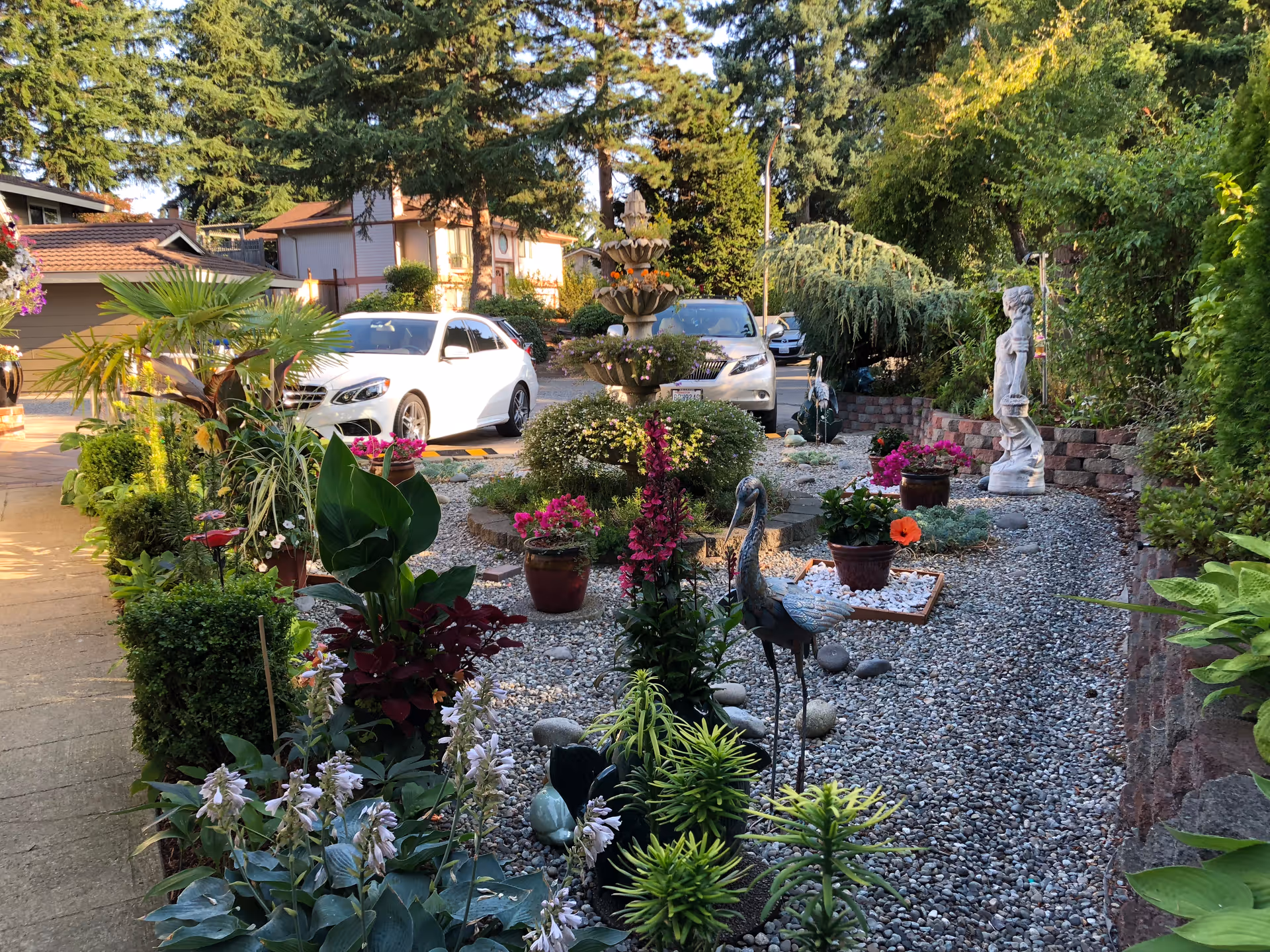 A landscaped outdoor garden area with various potted plants, flowers, and decorative statues including a bird and a human figure. There is a gravel path running through the garden, with a multi-tiered fountain in the center. In the background, there are parked cars and residential buildings surrounded by tall trees.