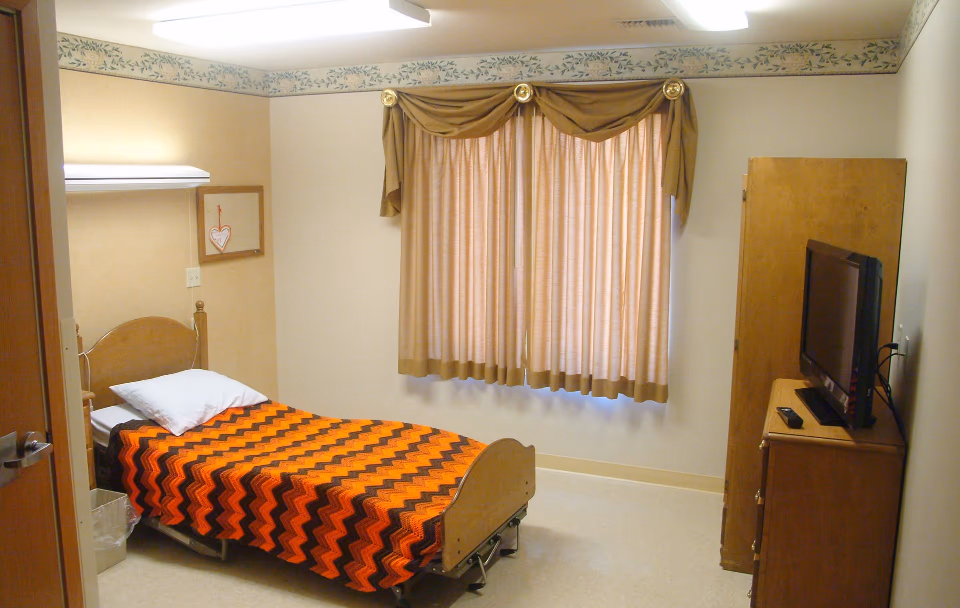 A small bedroom with a single bed covered by an orange and black zigzag patterned blanket. The bed has a white pillow and a wooden headboard. There is a window with beige curtains and a valance above it. A wooden dresser with a flat-screen TV and remote control is positioned against the right wall. The room has beige walls with a floral border near the ceiling and a closed wooden door on the left side.