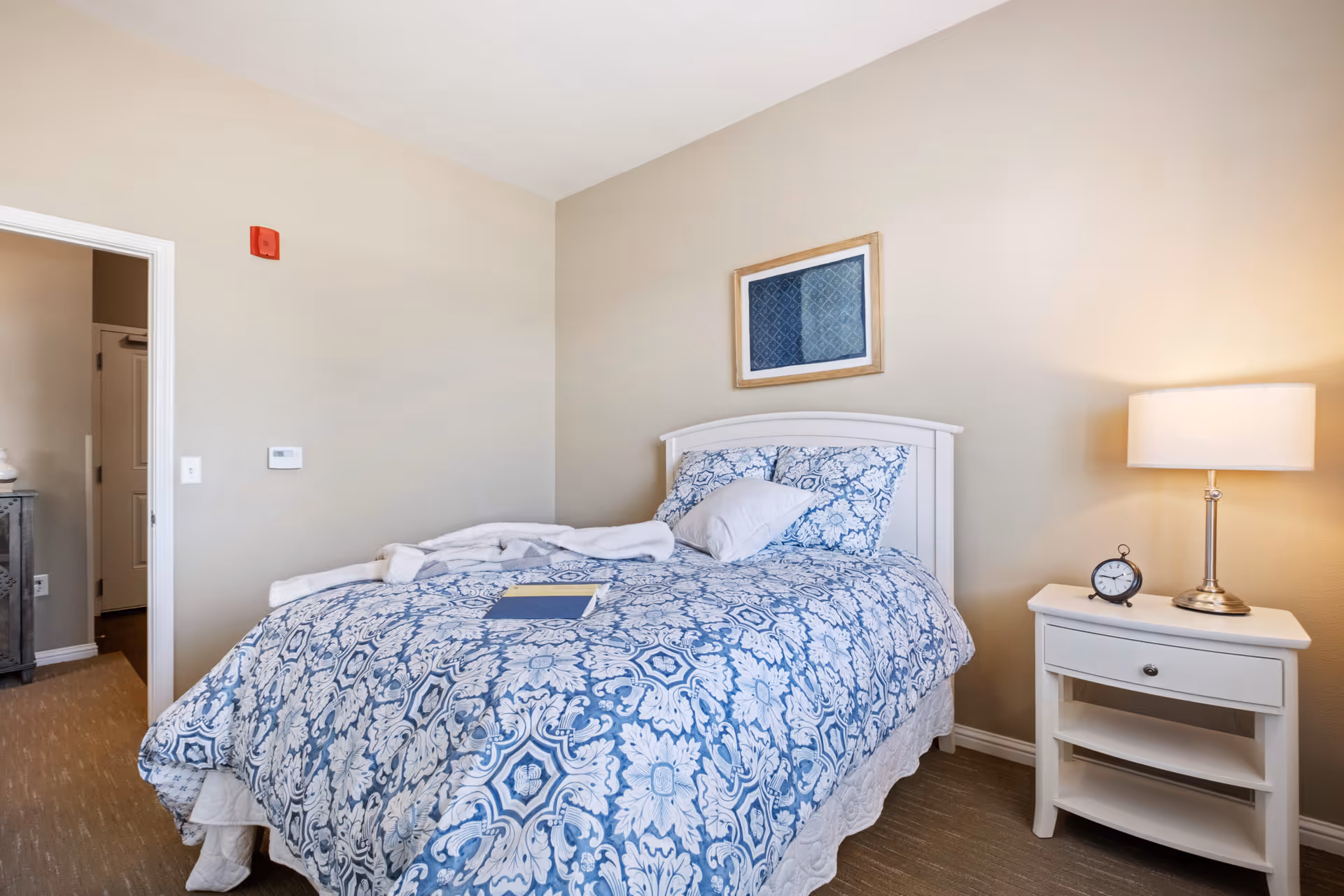 A cozy bedroom with a white wooden bed frame and a blue and white patterned comforter. There is a white pillow and a folded white blanket on the bed, along with a book. A white nightstand with a drawer and two shelves stands beside the bed, holding a small round clock and a lamp with a white shade. The walls are painted beige, and a framed blue artwork hangs above the bed. An open doorway leads to another room.